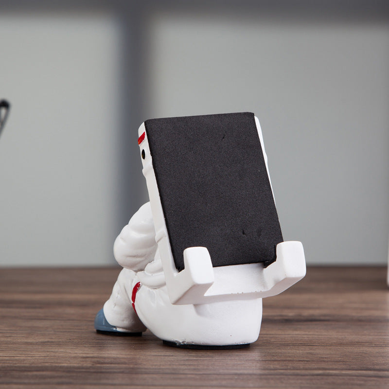 Decoratieve telefoonhouder in de vorm van een zittende astronaut, wit met zwarte helm en tablet in handen