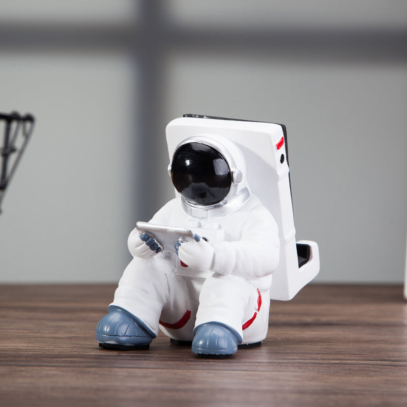 Decoratieve telefoonhouder in de vorm van een zittende astronaut, wit met zwarte helm en tablet in handen