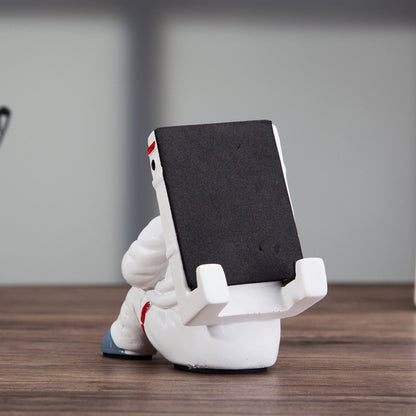 Decoratieve telefoonhouder in de vorm van een zittende astronaut, wit met zwarte helm en tablet in handen