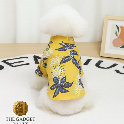Een witte, pluizige hond gefotografeerd van achteren in een geel Hawaiian-stijl hondenshirt met tropische print, zittend op een wit vloerkleed met subtiel The Gadget Vault-logo linksonder.
