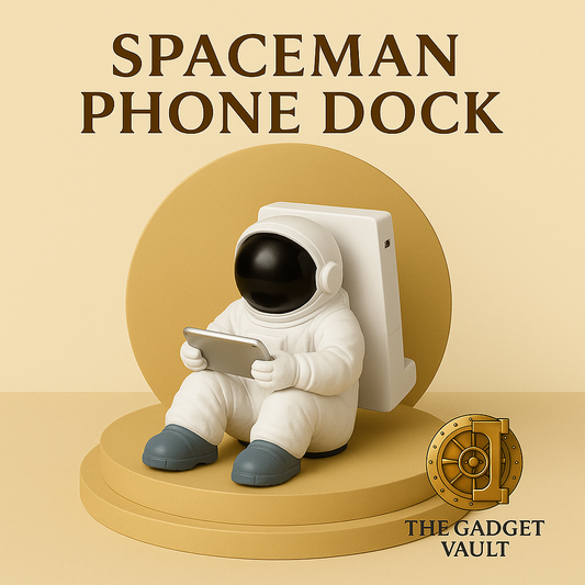 Decoratieve telefoonhouder in de vorm van een zittende astronaut, wit met zwarte helm en tablet in handen, geplaatst op een goud-beige achtergrond met het logo van The Gadget Vault rechtsonder.